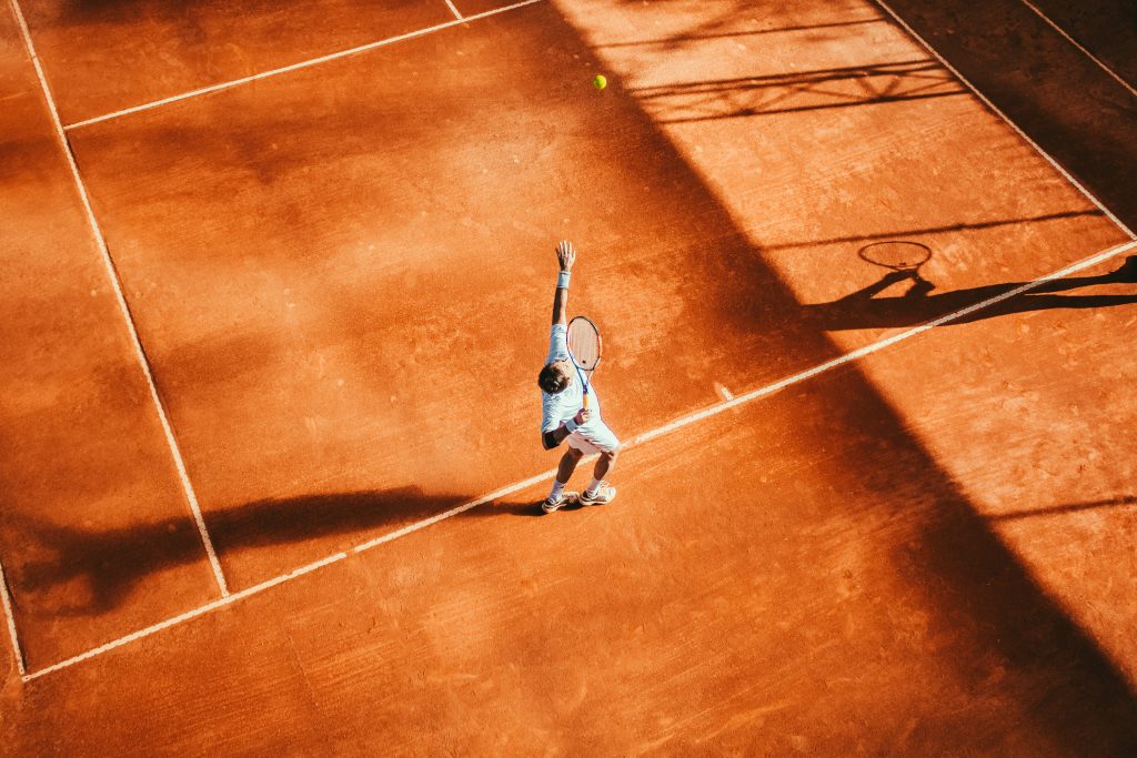 tennis athlete mid-serve on orange court in hot weather
